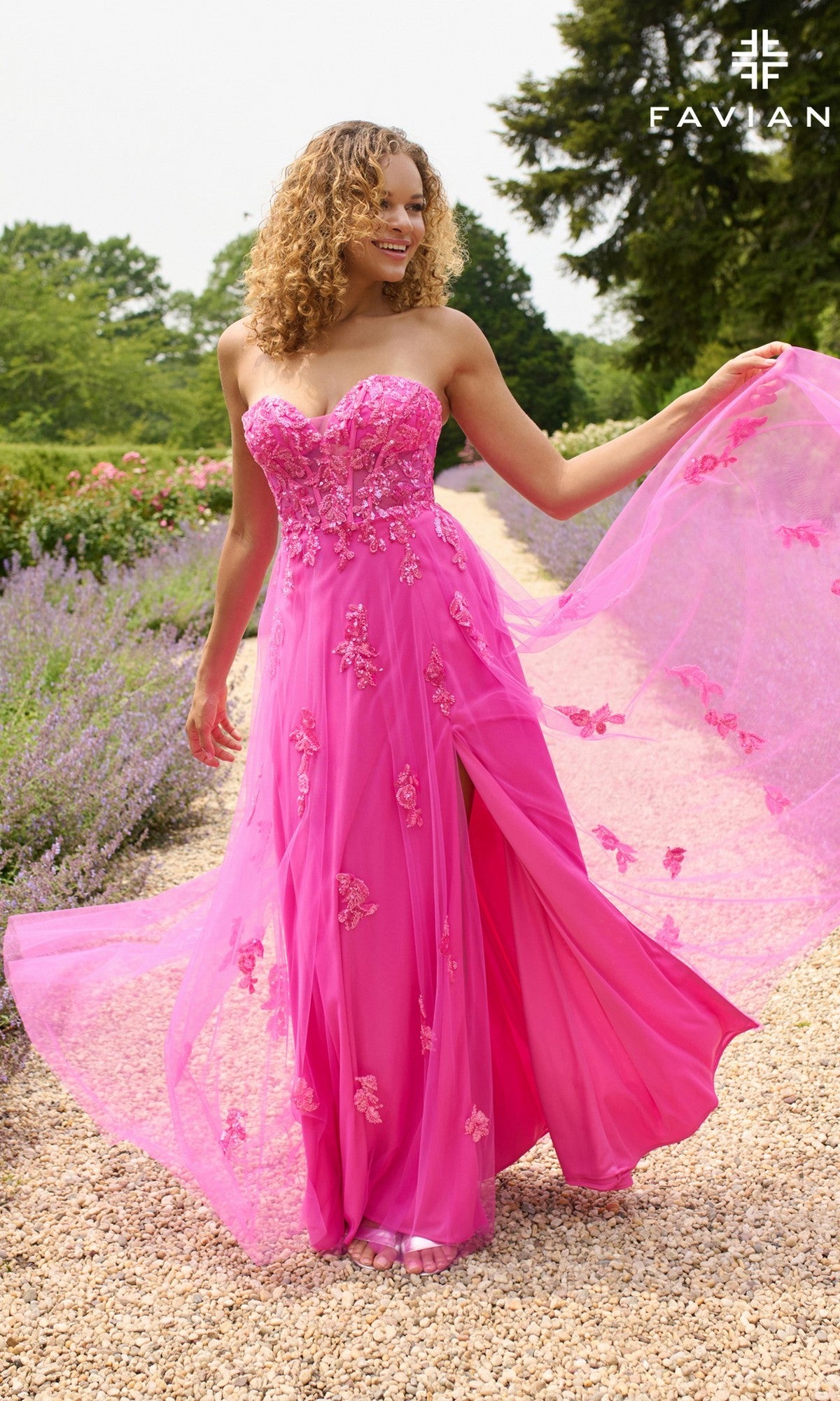 A woman wearing the Faviana 11474 long prom dress, featuring a strapless pink design with floral embroidery, beaded appliqué, and a thigh-high slit, stands on a gravel path in a garden.